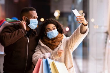 Karantina ve Salgın sırasında alışveriş. Mutlu Afro-Amerikan çifti tıbbi maske takıyor ve akşamları alışveriş merkezinin yakınında selfie çekiyorlar ellerinde alışveriş torbaları cep telefonlarının kameralarına gülümsüyorlar.