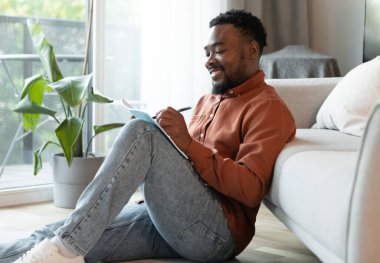 Cheerful Black Male Writer Writing And Taking Notes In Notebook Sitting On Floor At Home On Weekend. Creative Writing And Hobby Concept. Side View Shot