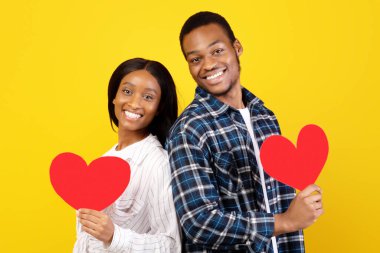 Sevgililer günün ve yıldönümün kutlu olsun. Gülümseyen binlerce yıllık Afro-Amerikan erkeği ve dişisi kırmızı kalpleri tutarlar ve kameraya bakarlar, sarı arka planda izole edilmiş, stüdyo çekimi, kopyalama alanı