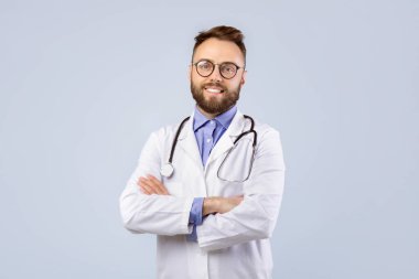 Laboratuvar önlüklü mutlu genç doktorun portresi kollarını kavuşturup gri stüdyonun arka planında kameraya bakıyor. Üniformalı, gülümseyen, pozitifti.