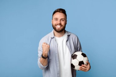 Futbol topu tutan neşeli genç adam, EVET jesti yapıyor, mavi stüdyo arka planında en sevdiği takımın kazanmasından mutlu. Dünya şampiyonası sırasında futbolcunun portresi. Spor ve eğlence.