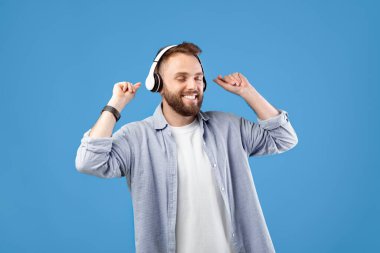 Kulaklıklı mutlu genç adam en sevdiği şarkıyla dans ediyor, mavi stüdyo arka planında müzik dinliyor. Neşeli milenyum adamı eğleniyor, güzel şarkı listesinden ve melodiden hoşlanıyor.