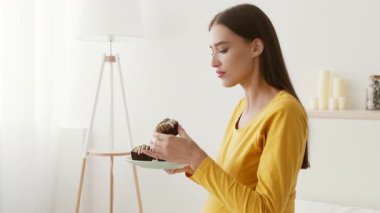 Hamile kadın aydınlık, dingin bir odada elinde bir tatlı tabağında duruyor. Sarı bir üst giyiyor ve sakin bir ortamın tadını çıkarıyor gibi görünüyor. Yumuşak ışıkla çevrili..