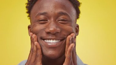 Neşeli bir Afrikalı genç Amerikalı adam yüzüne dokunurken geniş bir gülümseme sergiliyor, pozitiflik yayıyor. Canlı sarı arkaplan, bu canlı ortamda neşeli tavırlarını güçlendiriyor..