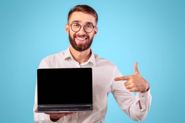 Pozitif genç girişimci mavi stüdyo arka planında web sitesi tasarımı için boş ekranlı dizüstü bilgisayarı işaret ediyor. Taşınabilir bilgisayarı olan mutlu milenyum işadamı. Model için alan.