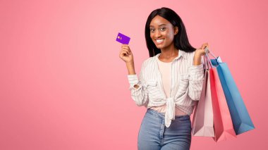 Kredi kartıyla hediye çantaları taşıyan ve pembe stüdyo arka planında gülümseyen güzel siyah bir kadın. Afrika kökenli Amerikalı kadın tatil hediyeleri için ücretsiz ödeme yapıyor, online indirimler sunuyor.