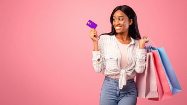 Hediye çantaları ve kredi kartıyla pembe stüdyo geçmişinde gülümseyen neşeli siyah kadın. Muhteşem Afro-Amerikan bayan tatil hediyeleri için online alışveriş yapıyor, büyük indirimler sunuyor.
