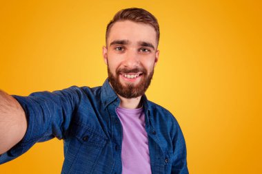 Yakışıklı beyaz adam selfie çekiyor ve turuncu stüdyo arka planında gülümsüyor. Pozitif milenyum adamı kendini kamerada fotoğraflıyor, kendi portresini yapıyor.