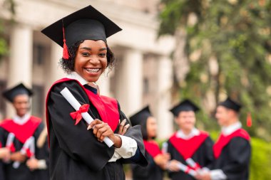 Mezuniyet kostümlü neşeli Afro-Amerikalı bayan diplomasını gösteriyor ve kameraya gülümsüyor. Üniversite kampüsündeki uluslararası öğrenci grubuna poz veren siyahi kız öğrenci.
