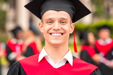 Mezuniyet kostümü giymiş yakışıklı bir öğrenci, siyah-kırmızı bornoz ve şapka giymiş, üniversite kampüsünde mezuniyet töreni yaparken kameraya neşeyle gülümsüyor, yakın plan portresi, fotokopi alanı.