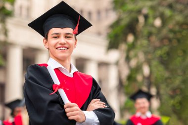 Mezuniyet cüppeli ve şapkalı mutlu genç adam kameraya gülümseyen ve üniversite kampüsünde sohbet eden çok kültürlü öğrencilerin başında duran.