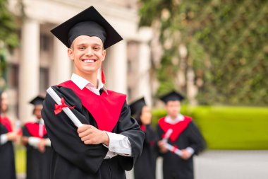 Mezuniyet kostümlü neşeli adam diplomasını gösteriyor ve kameraya gülümsüyor. Uluslararası öğrenci grubuna poz veriyor. Üniversite kampüsünde mezuniyeti kutluyorlar.