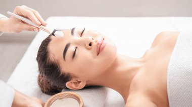 Güzellik Tedavisi. Profesyonel güzellik uzmanı spa salonundaki Asyalı kadına kilden maske takıyor. Masaj masasında uzanan çekici bir Koreli kadın. Yüz bakımının keyfini çıkarıyor.
