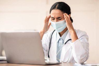 Kadın doktor, koruyucu yüz maskeli esmer kadın klinikte dizüstü bilgisayarla çalışırken baş ağrısı çekiyor, çalışma masasında gözleri kapalı oturuyor, şakaklarına dokunuyor, boşluğu kopyalıyor.