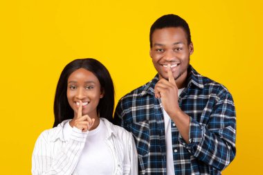Sır tutmak, haberleri paylaşmak, dudaklar şşşş hareketi ve özel bilgi. Gülümseyen esrarengiz Afro-Amerikan öğrencileri erkek ve kadın dudakları birbirine bastırıyor, sarı arka planda izole edilmiş.