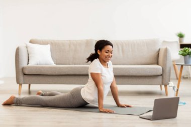 İyilik ve refah. Genç spor düşkünü Afrikalı Amerikalı kadın evde hatha yogası yapıyor, Kobra pozu veriyor, dizüstü bilgisayar kullanıyor. Spor ve sağlıklı yaşam tarzı kavramı.