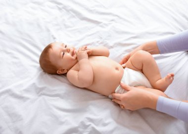 Tanımlanamayan anne gülümseyen küçük bebek bezini beyazla değiştiriyor. Sevimli sarışın çocuk, annesi onu giydirirken, parmaklarıyla oynarken, yakın plan, boş alan.