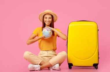 Elinde küre olan gülümseyen genç bir kadın, pembe arka planda sarı bavulun yanında oturuyor, seyahat etmek için yeni bir yer seçiyor, uzayı kopyalıyor. Neşeli bayan tatile hazırlanıyor, ülkeyi seçiyor.