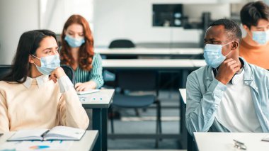 Modern eğitim, lise. Çok ırklı uluslararası sınıf arkadaşlarının portresi. Tek kullanımlık tıbbi yüz maskeleri takıyorlar ve konuşuyorlar, düşünceli insanlar sınıfta masada oturuyor, projeyi tartışıyorlar.