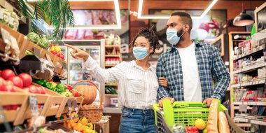 Süpermarket alıcıları. Afro-Amerikalı çift birlikte alışveriş yapıyor, sağlıklı gıda ürünleri alıyor, markette alışveriş arabalarıyla yürüyen yüz maskeleri takıyor. Müşteri ve Satış Konsepti