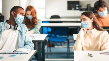 Eğitim ve Sağlık Konsepti. Çok kültürlü uluslararası sınıf arkadaşlarının portresi. Tek kullanımlık koruyucu maskeler takıyorlar, konuşuyorlar, sınıfta masada oturuyorlar, görevleri tartışıyorlar.