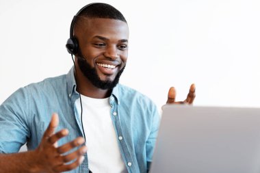 Video konferansı. Keyifli Afro-Amerikalı kulaklıklı bir adam iş yerindeki bilgisayarında web araması yapıyor, internet iletişiminin keyfini çıkarıyor, pozitif siyahi adam konuşuyor ve kameraya işaret veriyor.