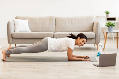 Çevrimiçi antrenman. Genç Afro-Amerikan Kadın Antrenman yapıyor, dizüstü bilgisayarda video izliyor, yatak odasında ya da oturma odasında plank yapıyor. Siyah Kadın Abs ve Vücut Eğitimi