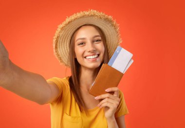 Happy young woman with passport and flight tickets taking selfie, yellow studio background