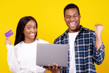 Alışveriş kolikleri ve internetten alışveriş. Covid-19 'da kendini izole etme. Heyecanlı genç Afro-Amerikan çifti duygularını ifade ediyor dizüstü bilgisayar ve kredi kartı, sarı arka planda izole edilmiş, stüdyo çekimi