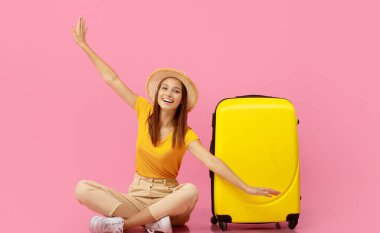 Uçağı taklit eden neşeli genç bayan gezgin, pembe stüdyo arka planında sarı bavulun üzerinde oturuyor, kopyalama alanı. Heyecanlı bayan tatil için hazırlanıyor, uçağı gösteriyor.