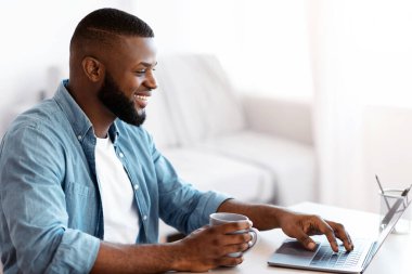 Telekomünikasyon konsepti. Evde dizüstü bilgisayarda çalışan gülümseyen siyahi adamın portresi bilgisayarla masada oturan pozitif Afrikalı adam, uzaktan çalışmanın tadını çıkartıyor, fotokopi ile yan görünümü var.