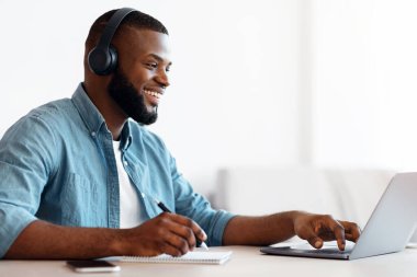 Çevrimiçi Eğitim. Kulaklıklı mutlu siyah adam internette çalışıyor, dizüstü bilgisayarda giyiniyor ve not alıyor, Afrikalı adam uzaktan öğrenmeyi seviyor, evde masa başında oturuyor,