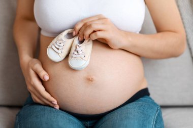 Hamile Bir Kadın 'ın Karnında Doğmamış Bebek İçin Küçük Ayakkabılar, Tanınmayan Anne Küçük Çizmeler Tutuyor ve Koltukta Otururken Göbeğini Kucaklıyor, Kesik Görüntü