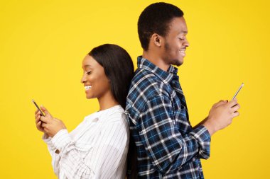 Sosyal ağ ve modern online ilişkiler. Gülümseyen Afrikalı genç Amerikalı erkek ve bayan sırt sırta durur, akıllı telefonlarda yazarlar, sarı arka planda izole edilmiş, stüdyo çekimi, profil, kopyalama alanı.