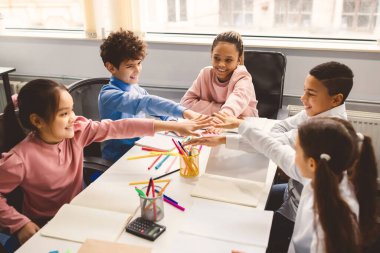 Çocuk Desteği Konsepti. Bir grup çok kültürlü, duygusal ortaokul öğrencisi modern sınıfta masa başında oturmuş el ele tutuşuyorlar, eğleniyorlar, ders çalışıyorlar veya oyun oynuyorlar.