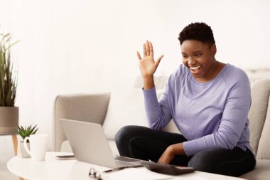 Çevrimiçi İletişim. Gülümseyen Afrikalı Afrikalı Amerikalı kadın dijital görüntülü konuşma yapıyor. İnterneti ve laptopu kullanarak ailesiyle Skype 'ta konuşuyor. Web kamerasına el sallıyor, oturma odasındaki koltukta oturuyor.