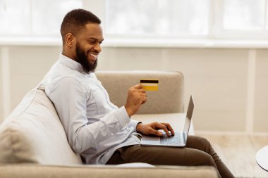Çevrimiçi alışveriş, perakende satış, alım. Gülümseyen sakallı siyah adam dizüstü bilgisayar kullanıyor ve internetten ürünler alıyor, bankayla veya kredi kartıyla ödeme yapıyor, oturma odasında kanepede oturuyor, bedava yer açıyor.