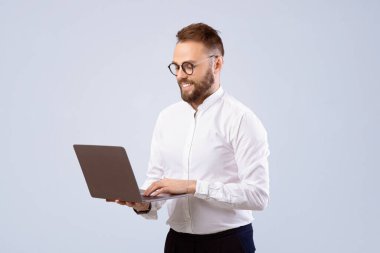 Ofisteki gülümseyen genç adam gri stüdyoda dizüstü bilgisayar kullanıyor. Millennial Freelancer uzaktan iş projesinde çalışıyor, online çalışıyor, web konferansı yapıyor.