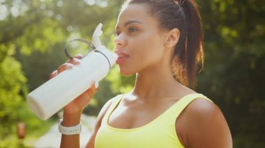 Genç bir Afrikalı Amerikalı kadın yemyeşil bir parkta koşuya ara verdi. Sağlıklı bir yaşam tarzını kucaklarken, güneşin altındaki yeşilliklerle çevrili bir şişeden suyu yudumluyor..