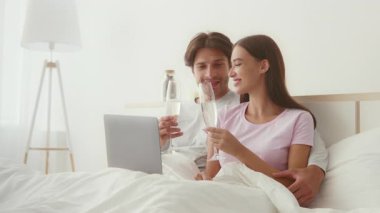 Sıcak bir çift, biri köpüklü bir içkiyi yudumlarken, yatağında dizüstü bilgisayarını paylaşarak huzurlu bir sabahın tadını çıkarıyor. Rahat atmosferleri sıcak ve samimi bir ortam yaratıyor..