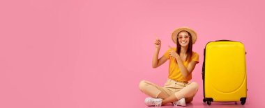 Yazlık kız valizin yanında oturuyor, pembe stüdyo arkaplanını işaret ediyor, kopyalama alanı.