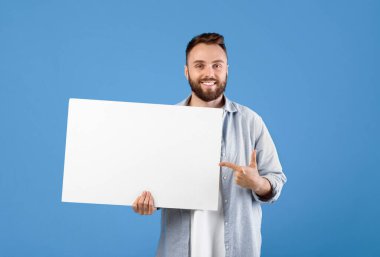 Reklam için boş yer. Mavi stüdyo arka planındaki boş beyaz tahtayı gösteren mutlu beyaz adam tasarım için bir model. Boş reklam panosu şablonuyla milenyum sakallı adam.