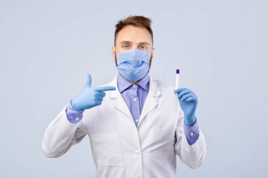Koruyucu maske ve eldiven takan erkek doktor gri stüdyo arka planındaki Coronavirus PCR test tüpünü işaret ediyor. Doktor nazal ve oral covid-19 tanı prosedürü için ilaç tutuyor.