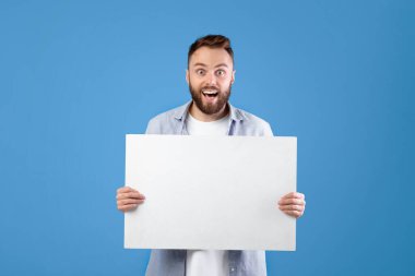 Heyecanlı genç adam elinde mavi stüdyo arka planında fotokopi alanı olan boş bir poster tutuyor. Mutlu milenyum adamı reklam tasarımınız için boş afiş gösteriyor.