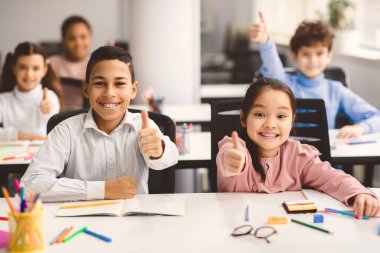 Eğitim Konsepti. Çeşitli mutlu, gülümseyen uluslararası sınıf arkadaşları sınıfta oturmuş işaret işareti gösteriyorlar. Kız ve oğlan okulda ders çalışmaktan zevk alıyorlar.