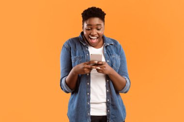 Sosyal medyada cep telefonu mesajları kullanan Afrikalı Amerikalı Heyecanlı Kadın ya da Sarı Stüdyo Arkaplanının Üzerinde Durarak Mobil Oyun oynamak. Akıllı Telefon Konseptiniz için Harika Cep Telefonu Uygulaması