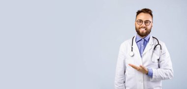Üniformalı neşeli erkek doktor gri stüdyo arka planındaki boşluğa bakıyor, panorama. Pratisyen hekim kameraya bakıyor ve tıbbi hizmetler sunuyor, afiş tasarımı