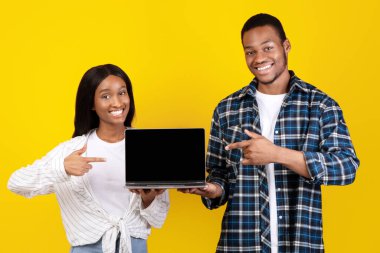 İnternet sitesi reklamı, çevrimiçi uygulama ve COVID-19 tecrit sırasında kendini izole etme. Neşeli genç Afrikalı Amerikalı karı koca dizüstü bilgisayarı boş ekranla tutuyorlar ve sarı arka planda parmaklarıyla gösteriyorlar.
