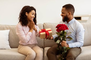 Sürpriz hazırlıyorum. Heyecanlı güzel Afrikalı Amerikalı kız arkadaşı mutlu erkek arkadaşından hediye kutusu ve bir buket kırmızı gül alıyor. Gülümseyen genç siyah çift evdeki kanepede oturuyor.