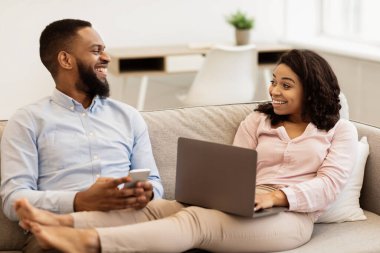 Hafta sonu tatili. Mutlu Milenyum Siyah Çifti 'nin portresi koltukta dinleniyor. Smarthone 'da Erkek Sohbeti, Kadın Laptop Kullanıyor. Aile birlikte çalışıyor ve internette geziniyor, konuşuyor ve gülüyor.